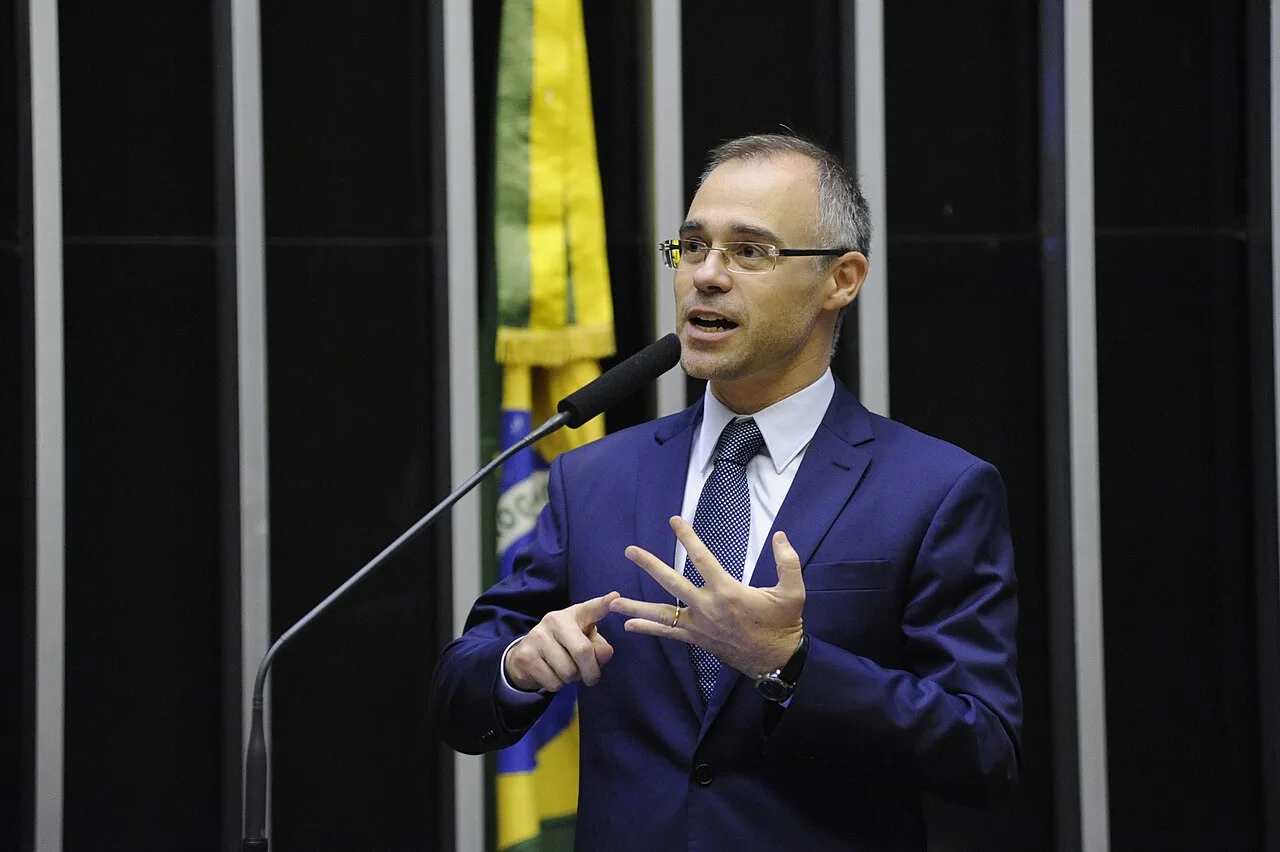 André Mendonça de óculos e terno escuro, gesticulando ao microfone com a bandeira do Brasil ao fundo