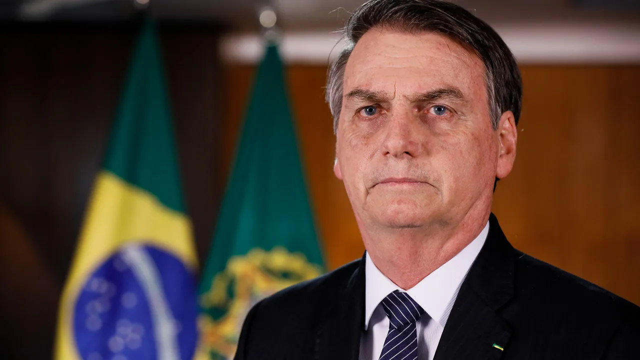 Jair Bolsonaro de terno e gravata em retrato oficial, com bandeiras do Brasil ao fundo
